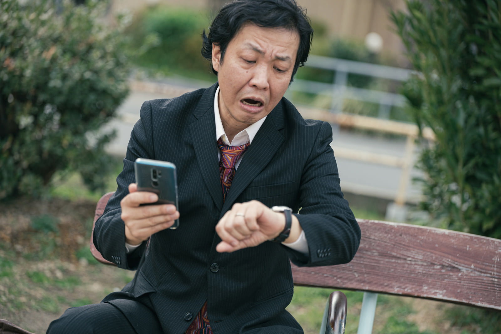 公園のベンチに座り慌ててスマートフォンで時間確認するスーツ姿の男性［モデル：のせさん］