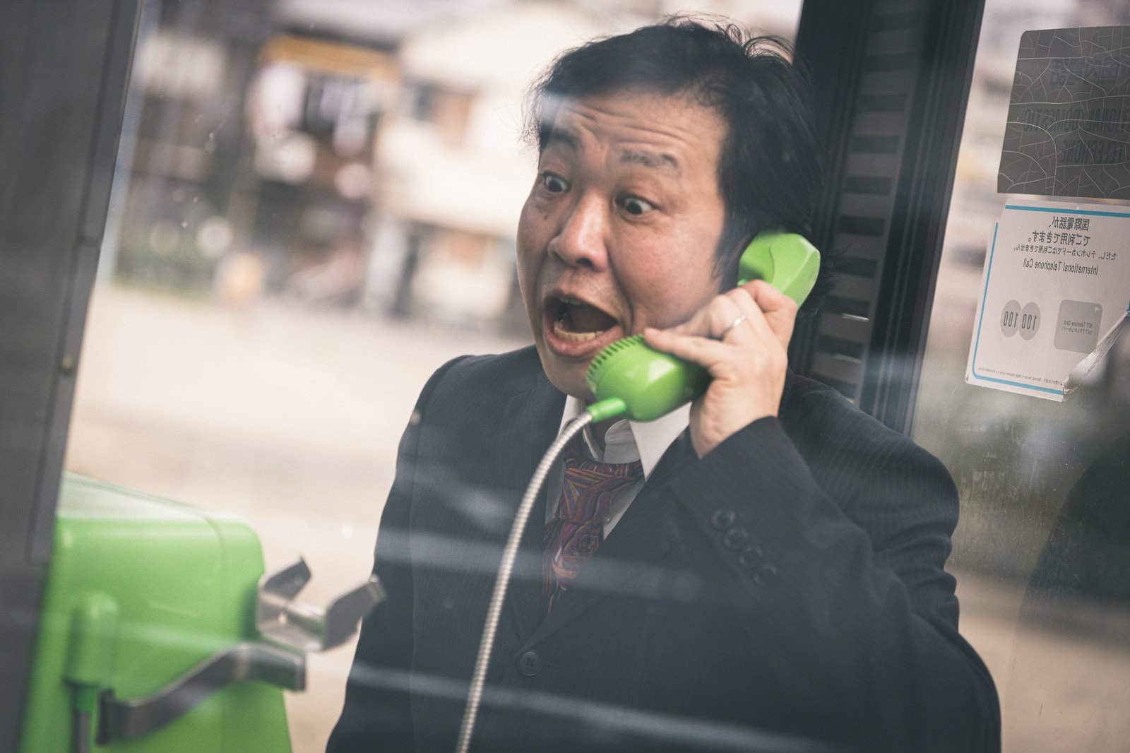 緑色の公衆電話で真剣な表情で通話するスーツ姿の男性［モデル：のせさん］
