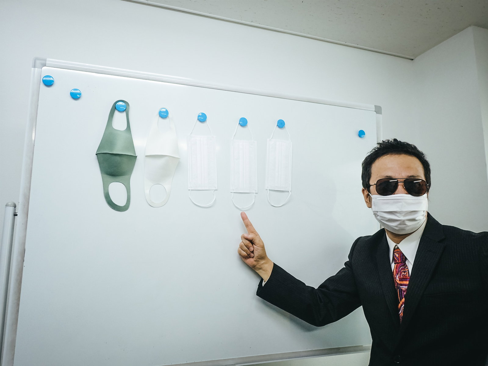 サングラスをかけたスーツ姿の男性がホワイトボードでマスクの説明をしている［モデル：のせさん］
