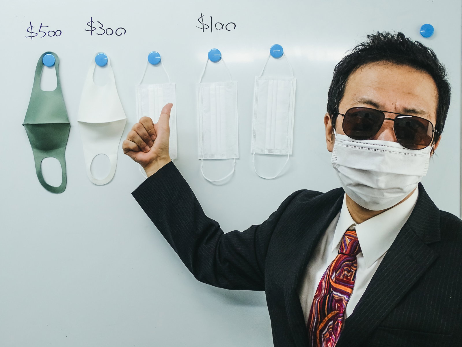 マスクとサングラスで顔を隠した黒いスーツの男性が指を立てている［モデル：のせさん］