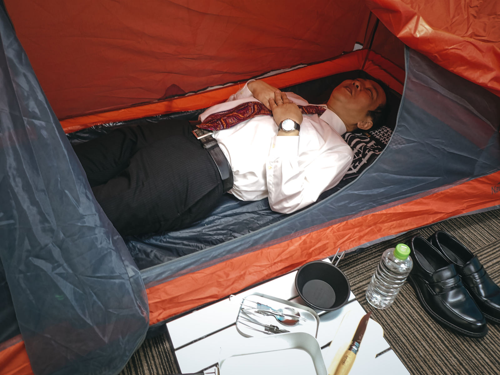 オレンジ色のテントの中でスーツ姿で横になって寝る会社員男性［モデル：のせさん］