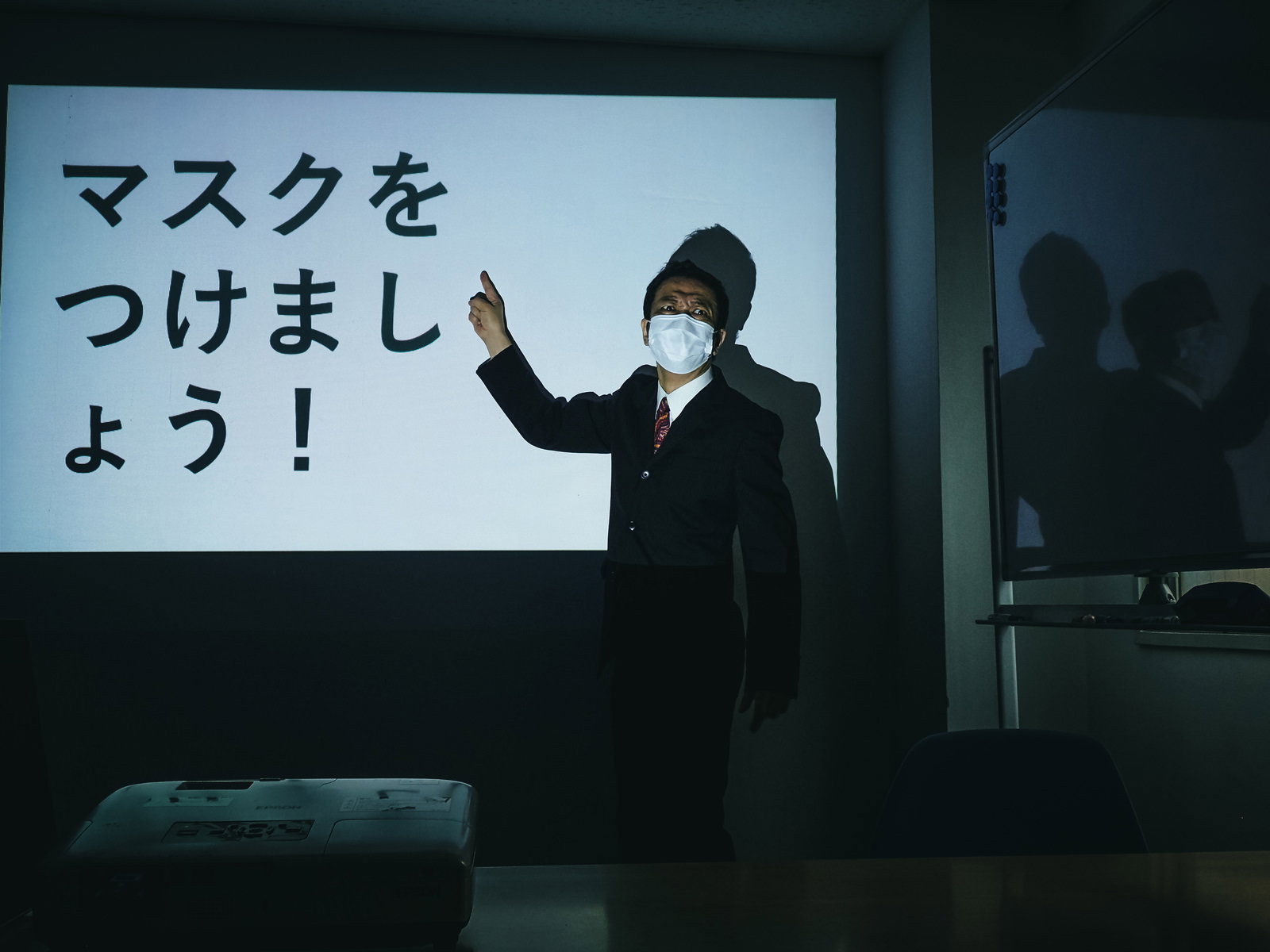 会議室でプロジェクターの前に立ち、白いマスクを着けて右手を指差しで上げている黒いスーツの男性［モデル：のせさん］