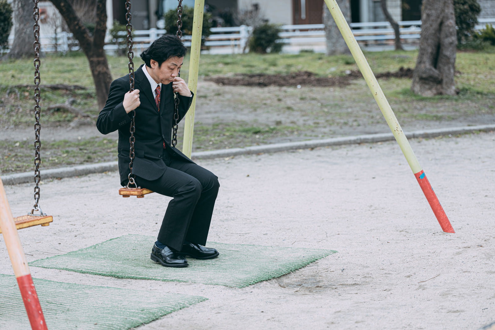 公園のブランコに座るスーツ姿の男性［モデル：のせさん］