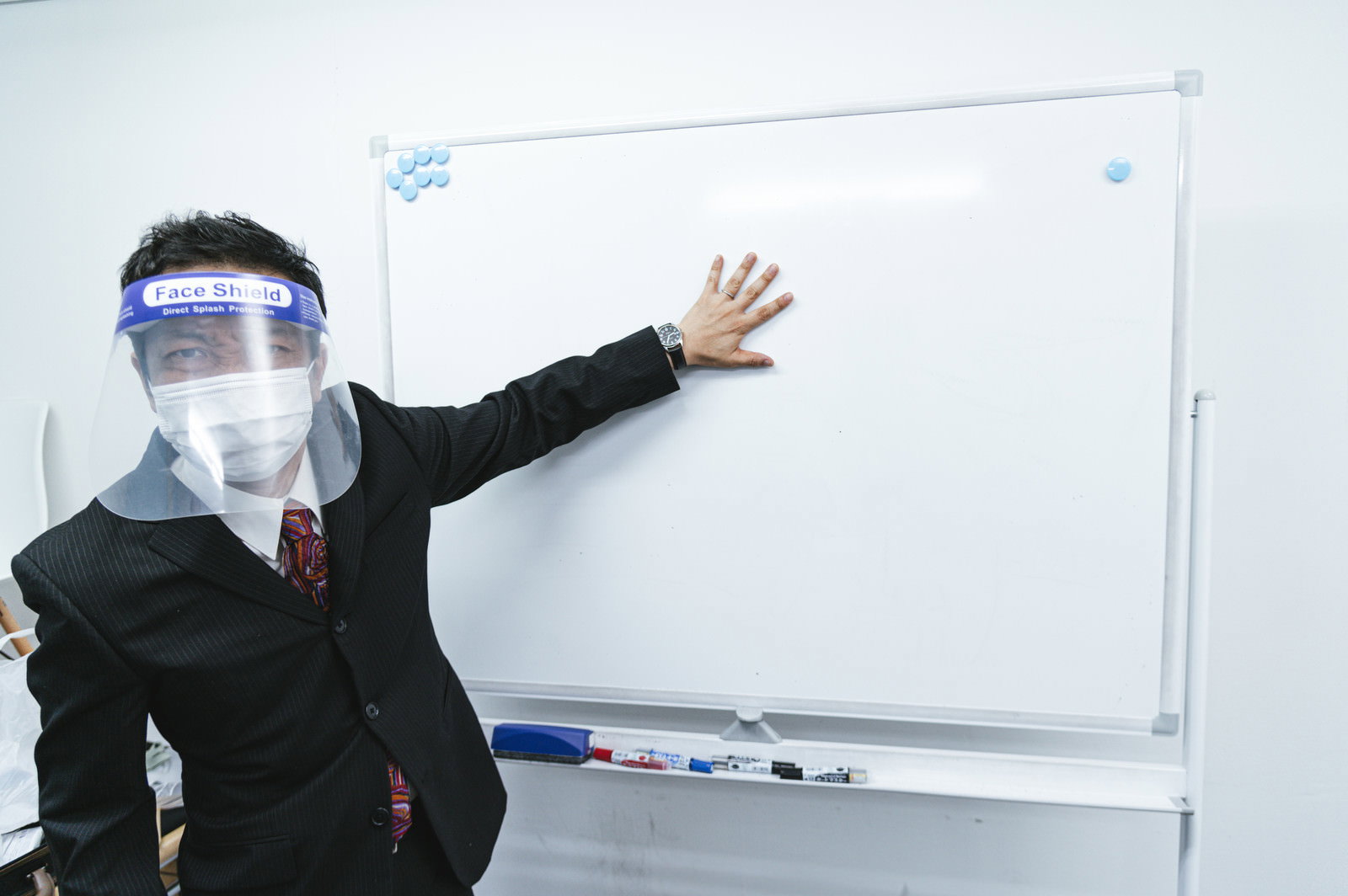 フェイスシールドを装着してホワイトボードの前でプレゼンする会社員［モデル：のせさん］