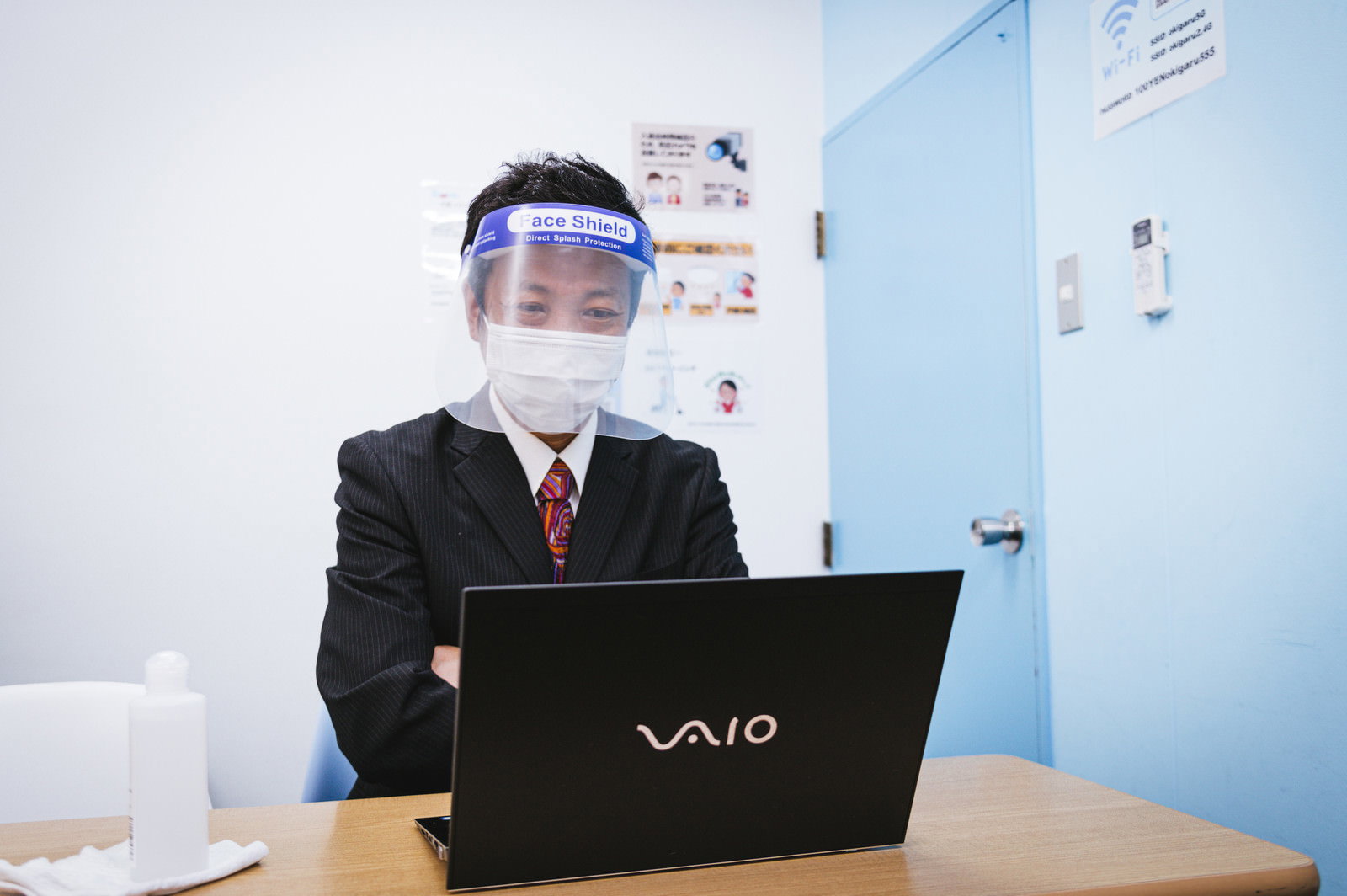 フェイスシールドとマスクを二重に着用してノートパソコンで作業している会社員［モデル：のせさん］