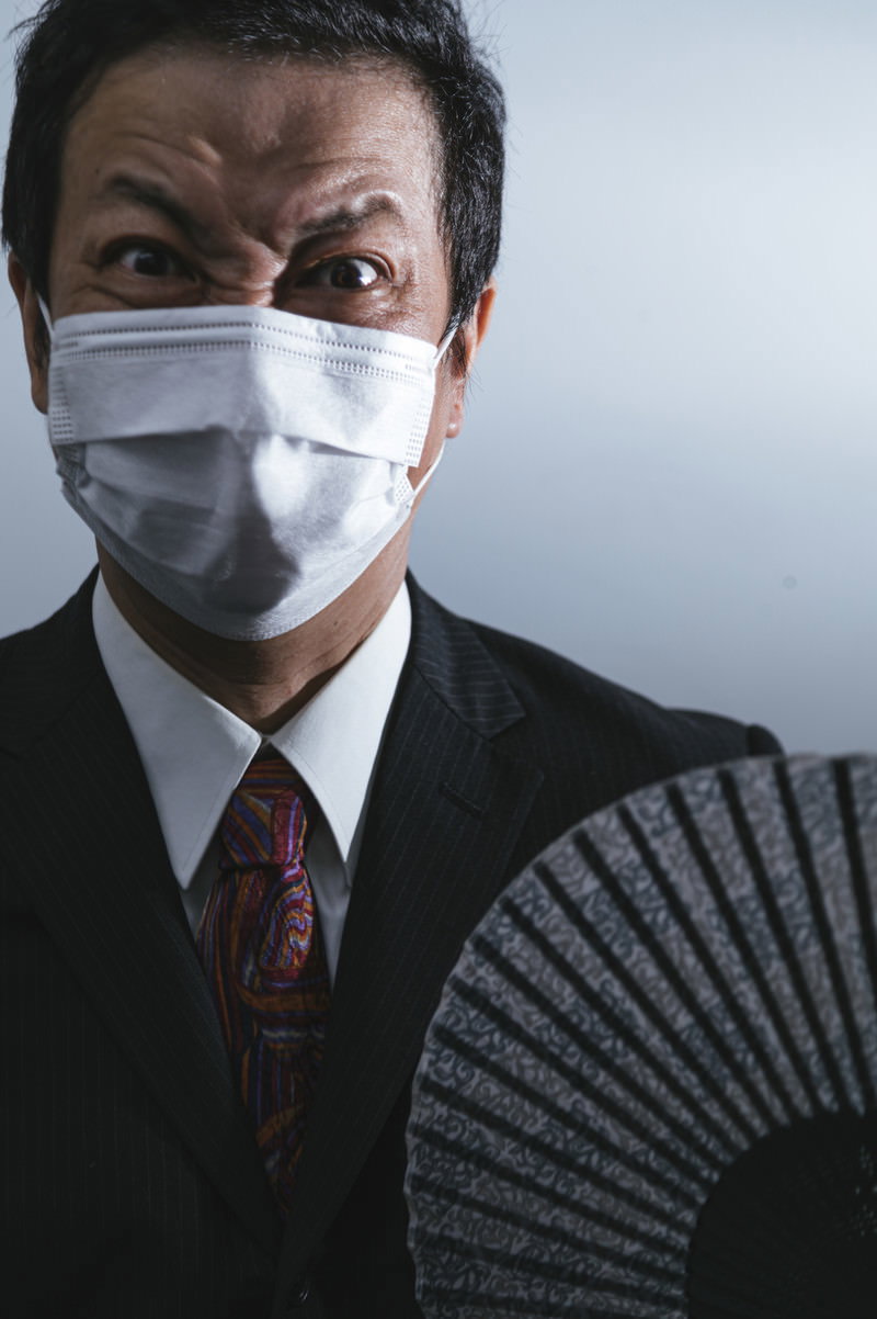 マスクを着用し扇子を持って怒りの表情を見せるスーツ姿の男性［モデル：のせさん］