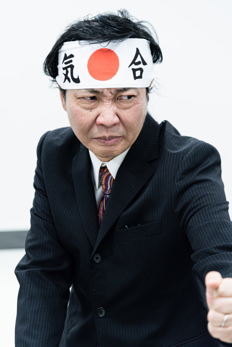 白い鉢巻きに赤い日の丸を付けて気合いを入れた表情の黒いスーツの男性［モデル：のせさん］