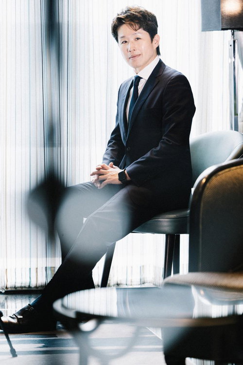 窓際のオフィスチェアに座り微笑むスーツ姿のM&Aアドバイザー