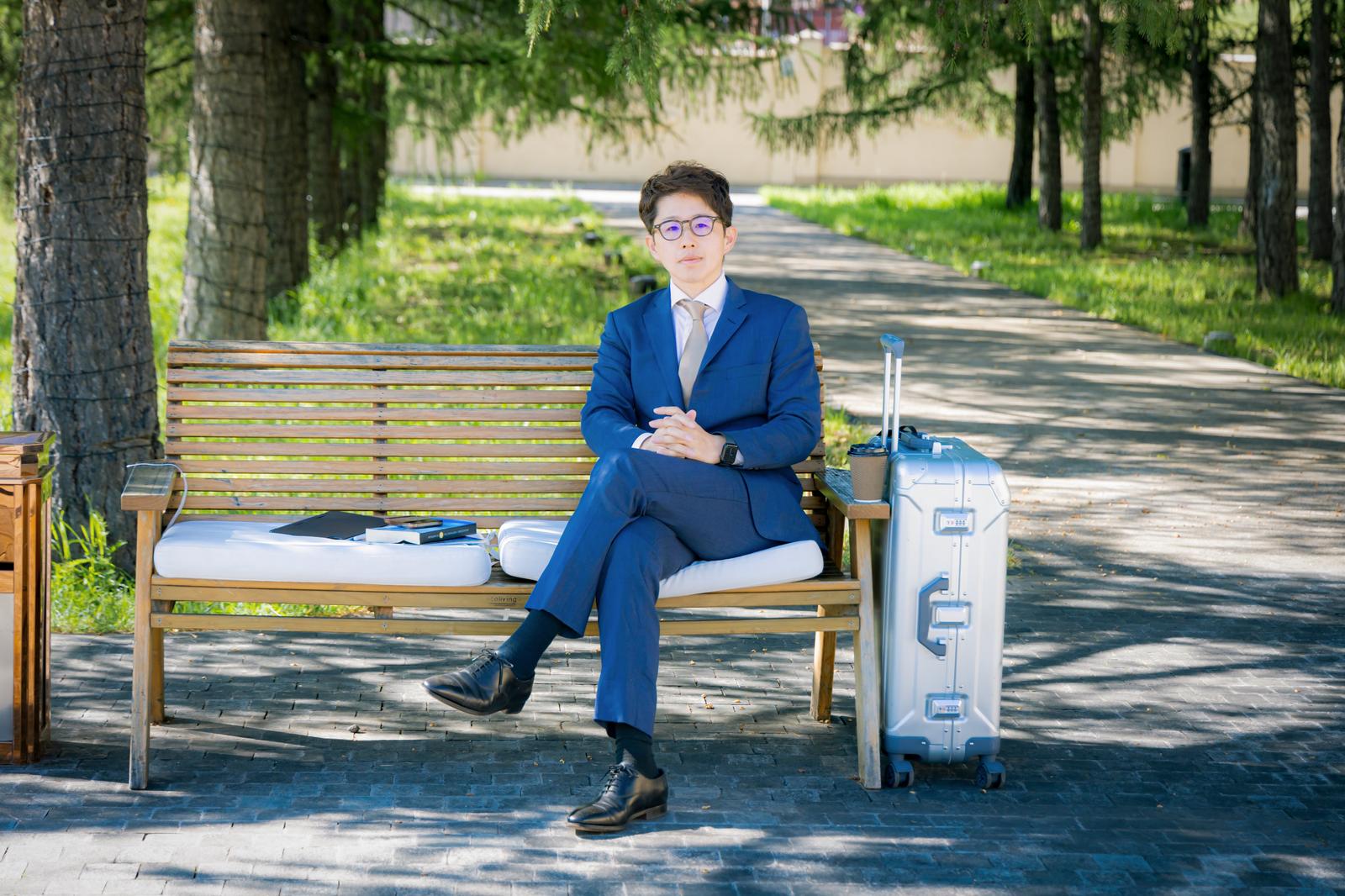 公園のベンチで待つビジネスマン、スーツ姿でキャリーバッグを横に置いている［モデル：野崎繁裕］
