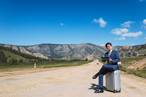 モンゴルの山道でキャリーケースに座ってビジネスをこなすスーツ姿の男性