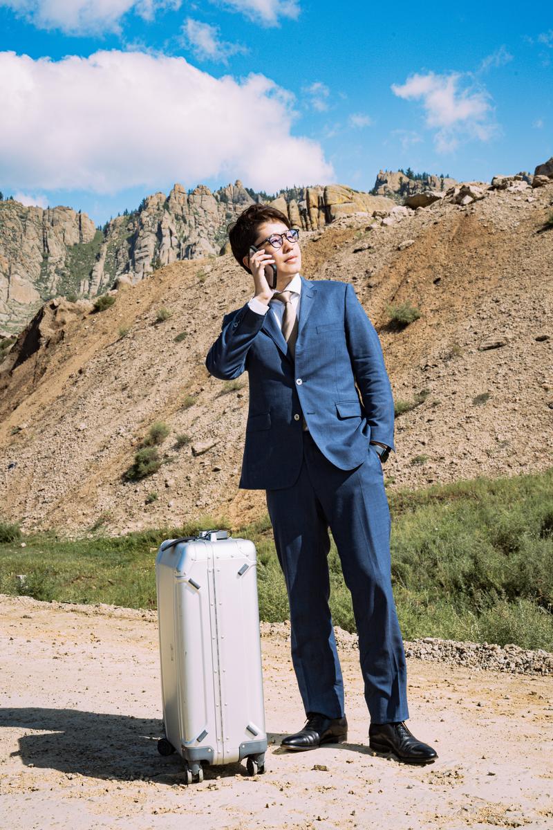 モンゴルの岩山の麓で白いキャリーバッグを持ちながらスマートフォンで通話する紺色スーツの男性［モデル：野崎繁裕］
