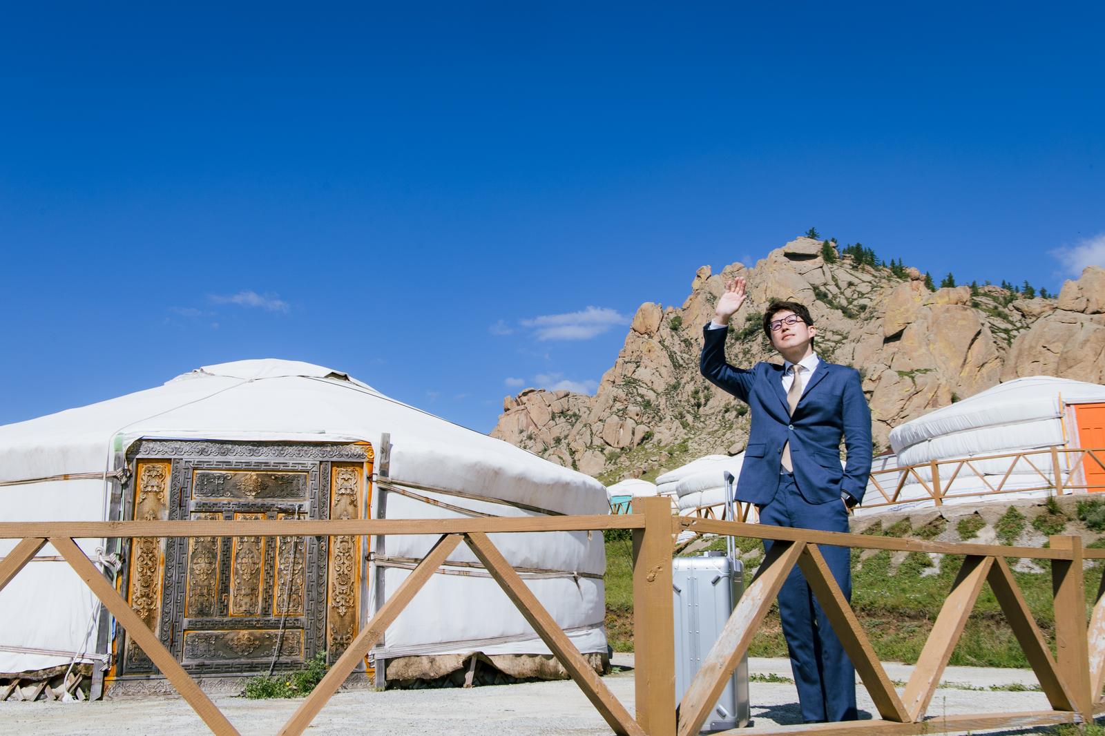 モンゴルのゲル前で紺色のスーツを着た男性が手を上げている［モデル：野崎繁裕］