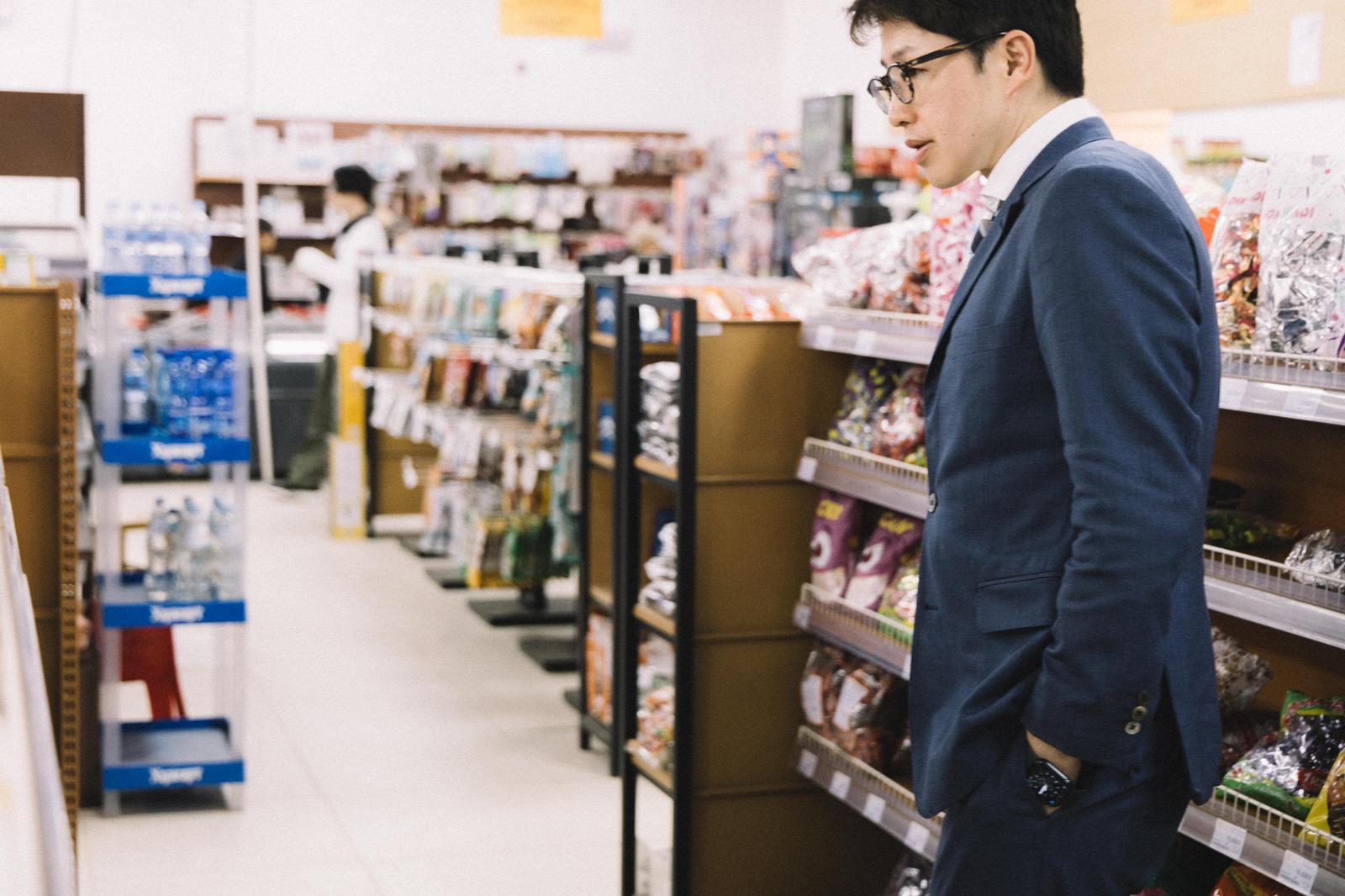 スーパーの売り場で商品を確認する紺色スーツの眼鏡をかけた男性［モデル：野崎繁裕］