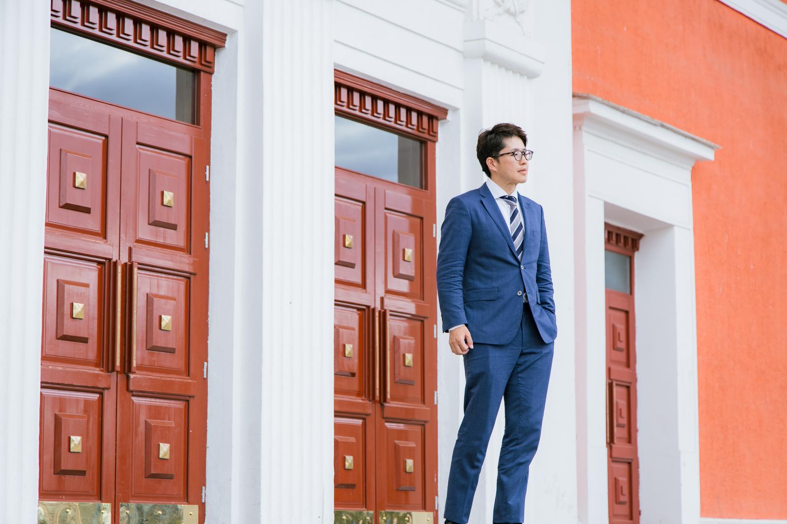 世界銀行の赤い扉の前に立つ、濃紺のスーツ姿のビジネスマン［モデル：野崎繁裕］