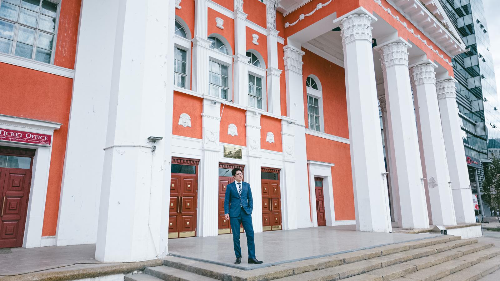 赤と白の建物の前で立つスーツ姿の男性［モデル：野崎繁裕］