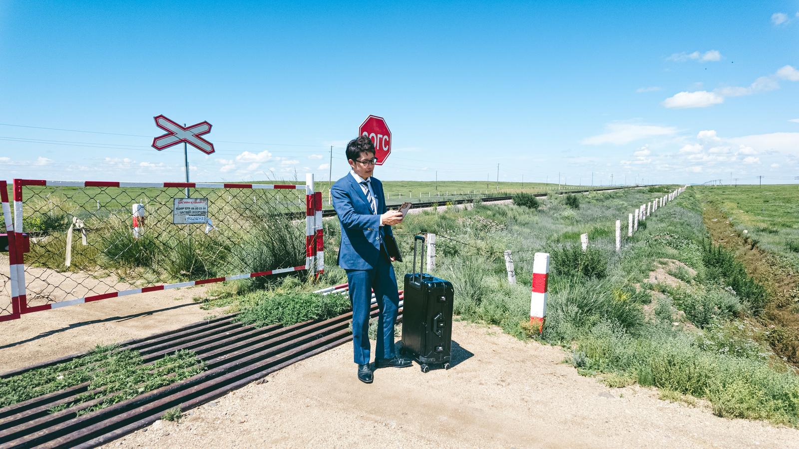 A man in a suit holding a black suitcase waiting for a train at a crossing in Mongolia