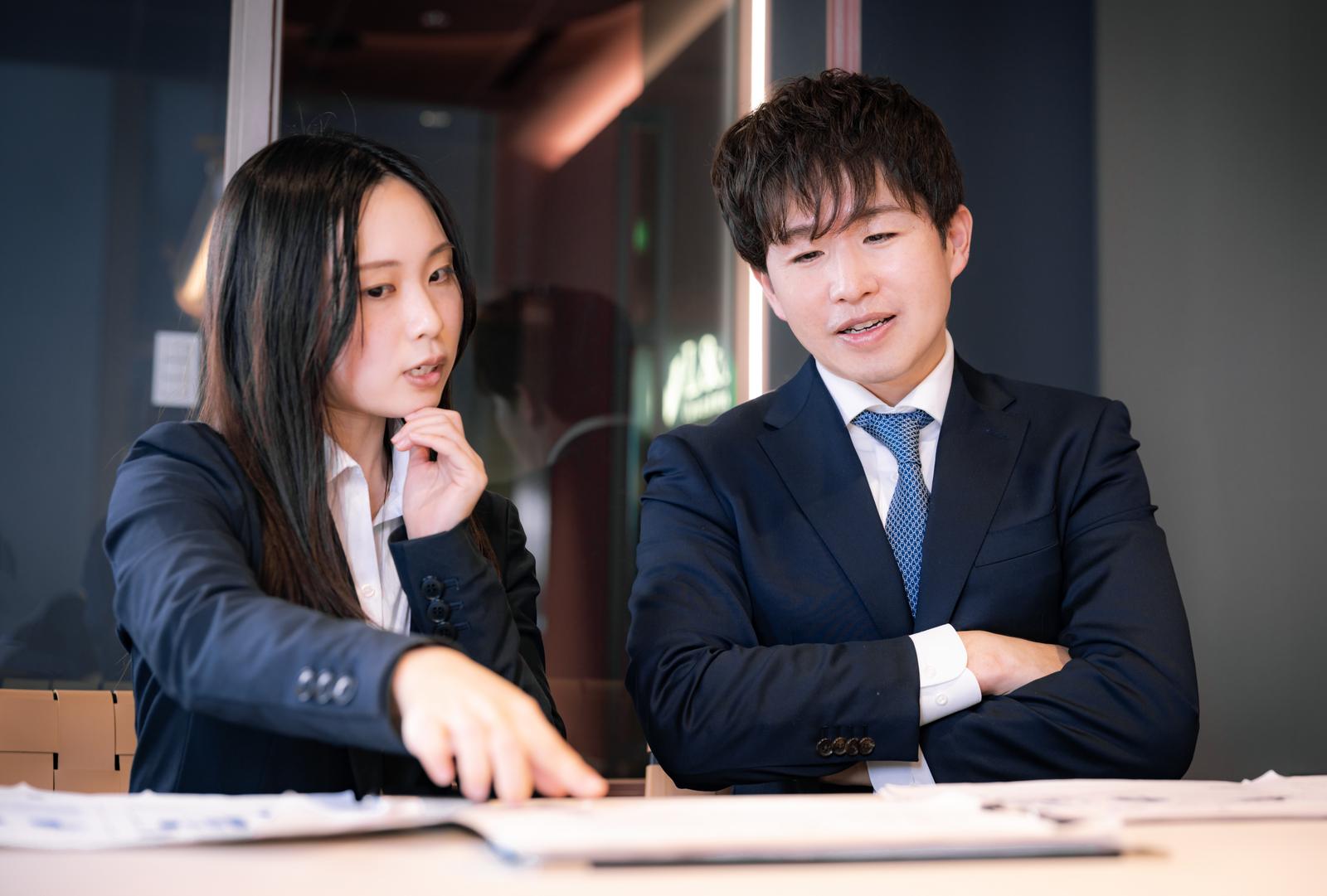 デスクで書類を見ながら相談するM&Aアドバイザーの男女［モデル：野崎繁裕 吉屋ゆり］