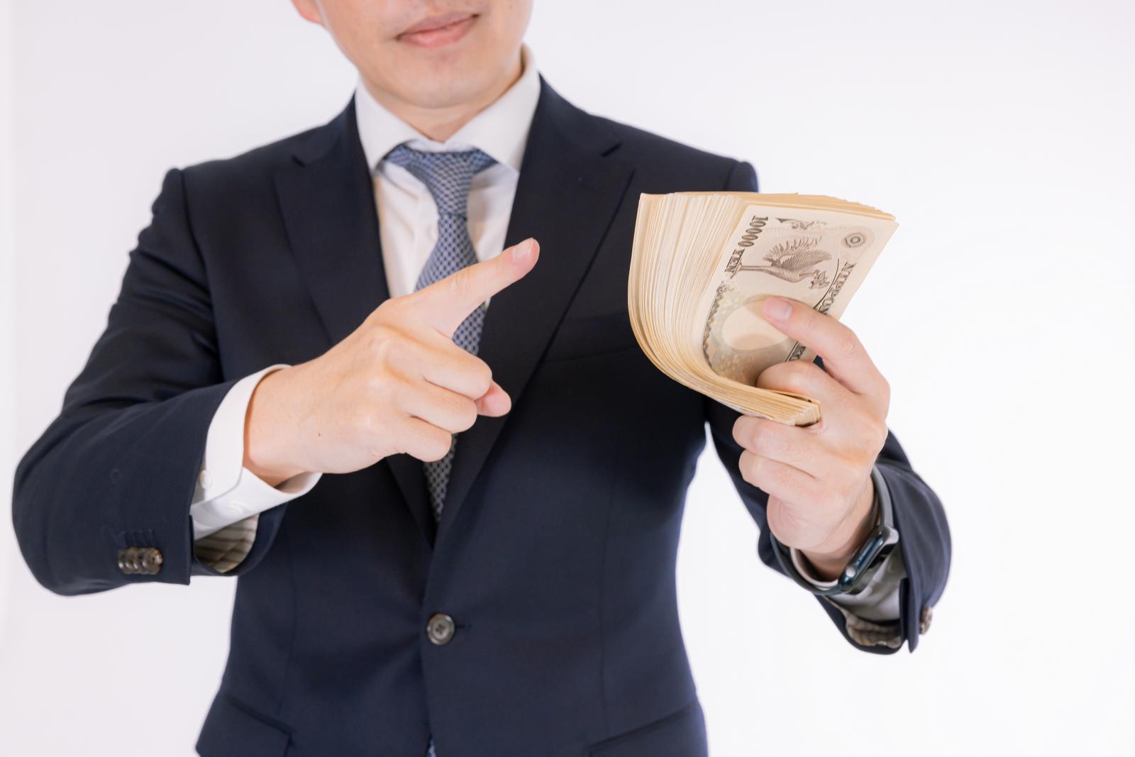 札束を指で示しながら説明する紺色スーツの男性ビジネスパーソン［モデル：野崎繁裕］
