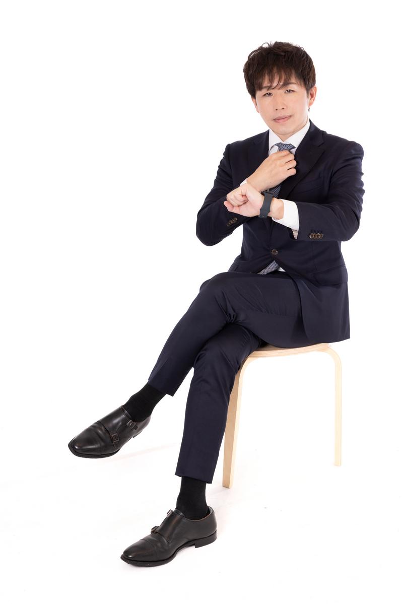 紺色スーツを着た男性がネクタイを調整しながら椅子に座っている［モデル：野崎繁裕］