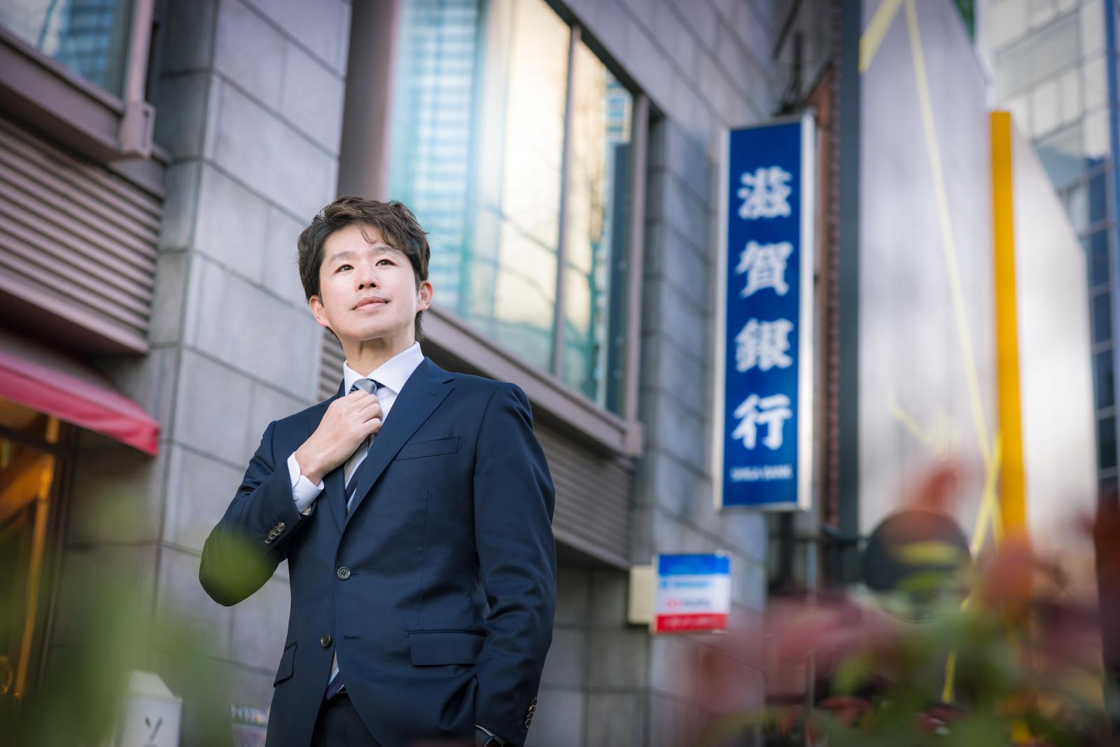 金融街で紺色のスーツを着てネクタイを整える男性M&Aアドバイザー［モデル：野崎繁裕］