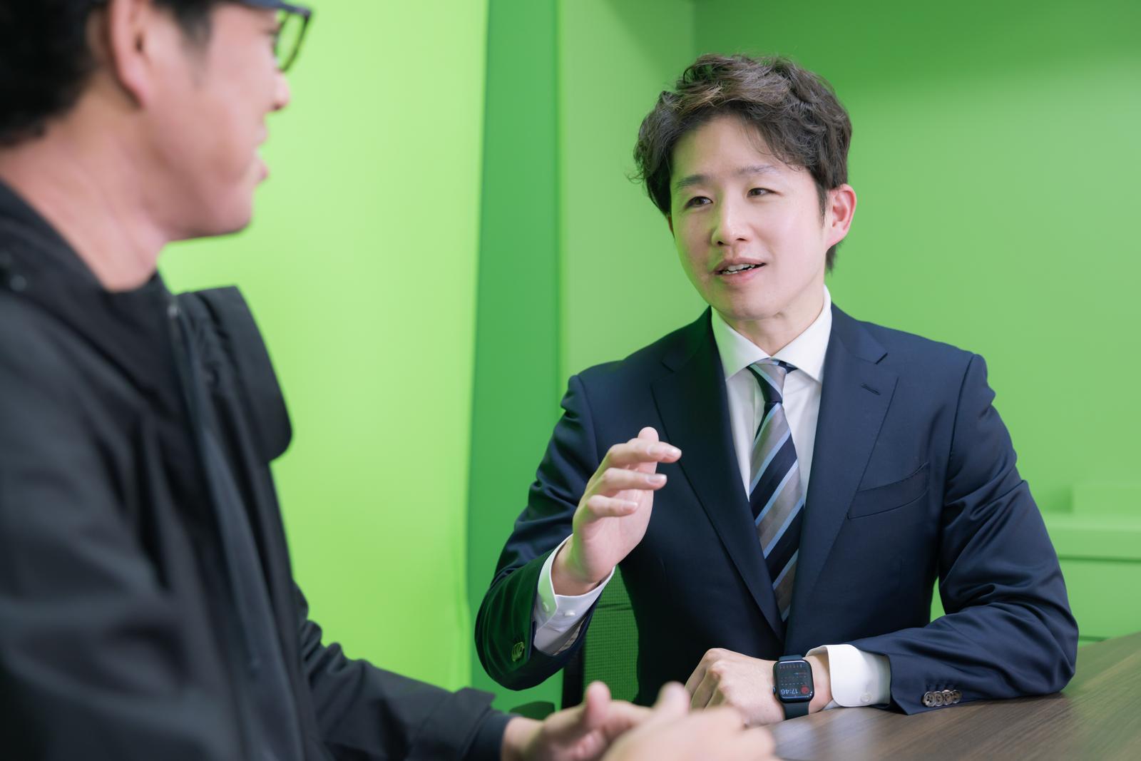 グリーンバックでインタビューするスーツ姿の男性［モデル：野崎繁裕］