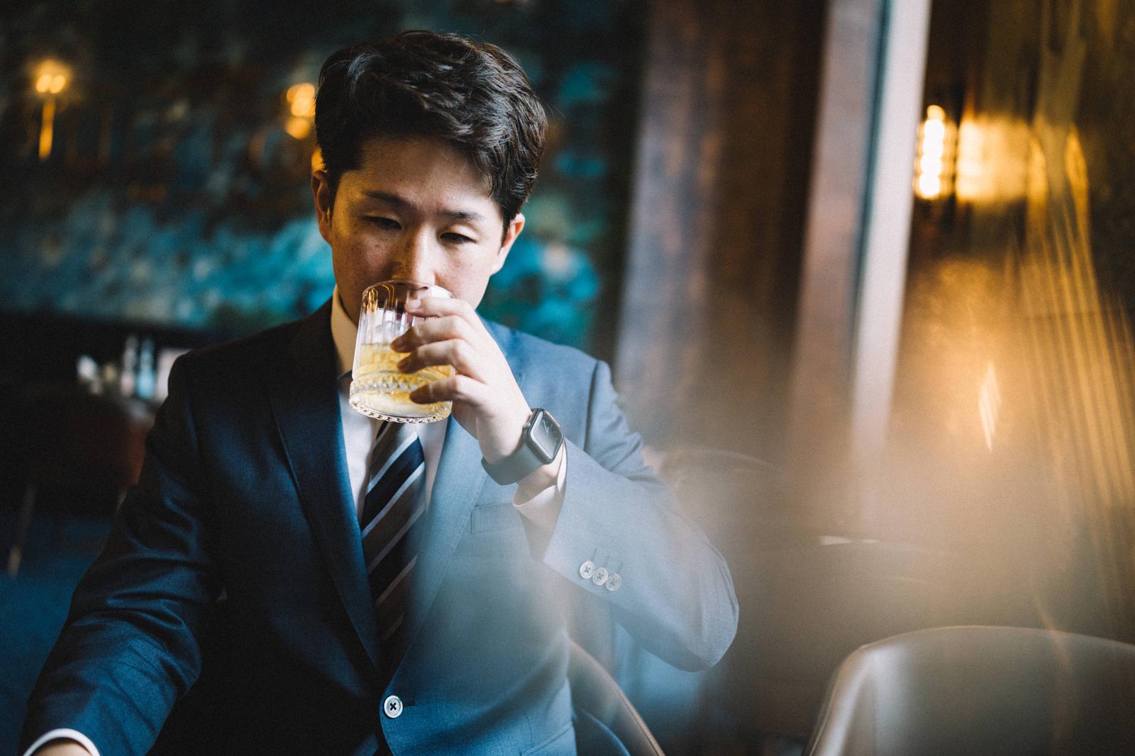 ウイスキーグラスを手に持つスーツ姿のビジネスマンがバーで飲酒している［モデル：野崎繁裕］