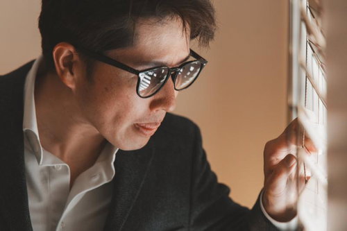 ブラインドの隙間から外を覗く眼鏡をかけた男性