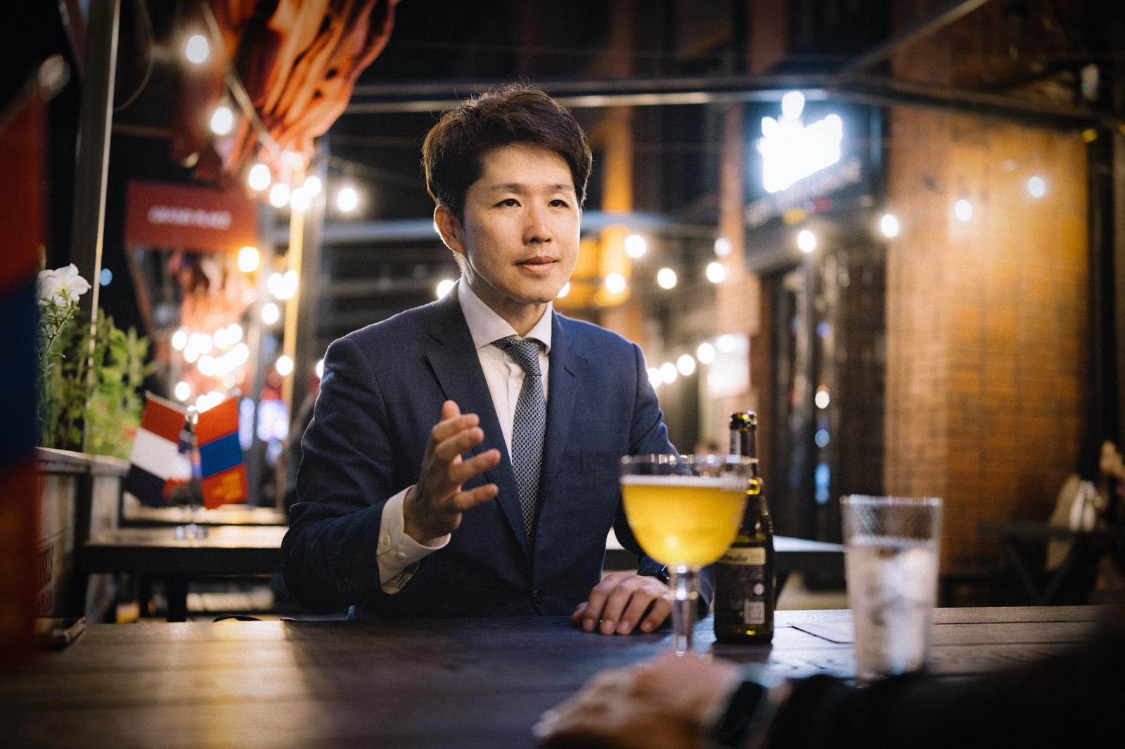 夜間の繁華街バーテラス席でビールを前に会話するスーツ姿のビジネスマン［モデル：野崎繁裕］
