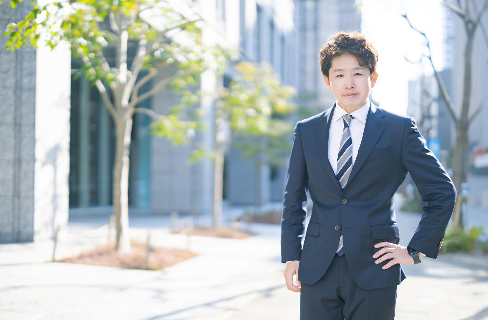 スーツ姿のM&Aアドバイザー男性がビジネス街に立つ［モデル：野崎繁裕］
