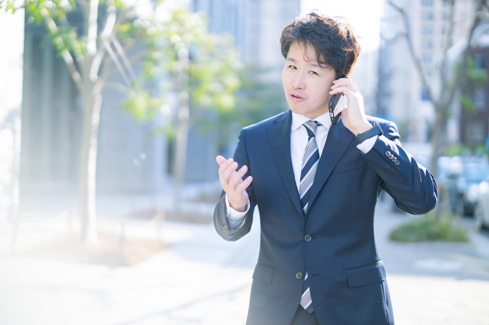 スーツを着た男性がスマートフォンで電話しながら屋外で説明しているビジネスシーン［モデル：野崎繁裕］