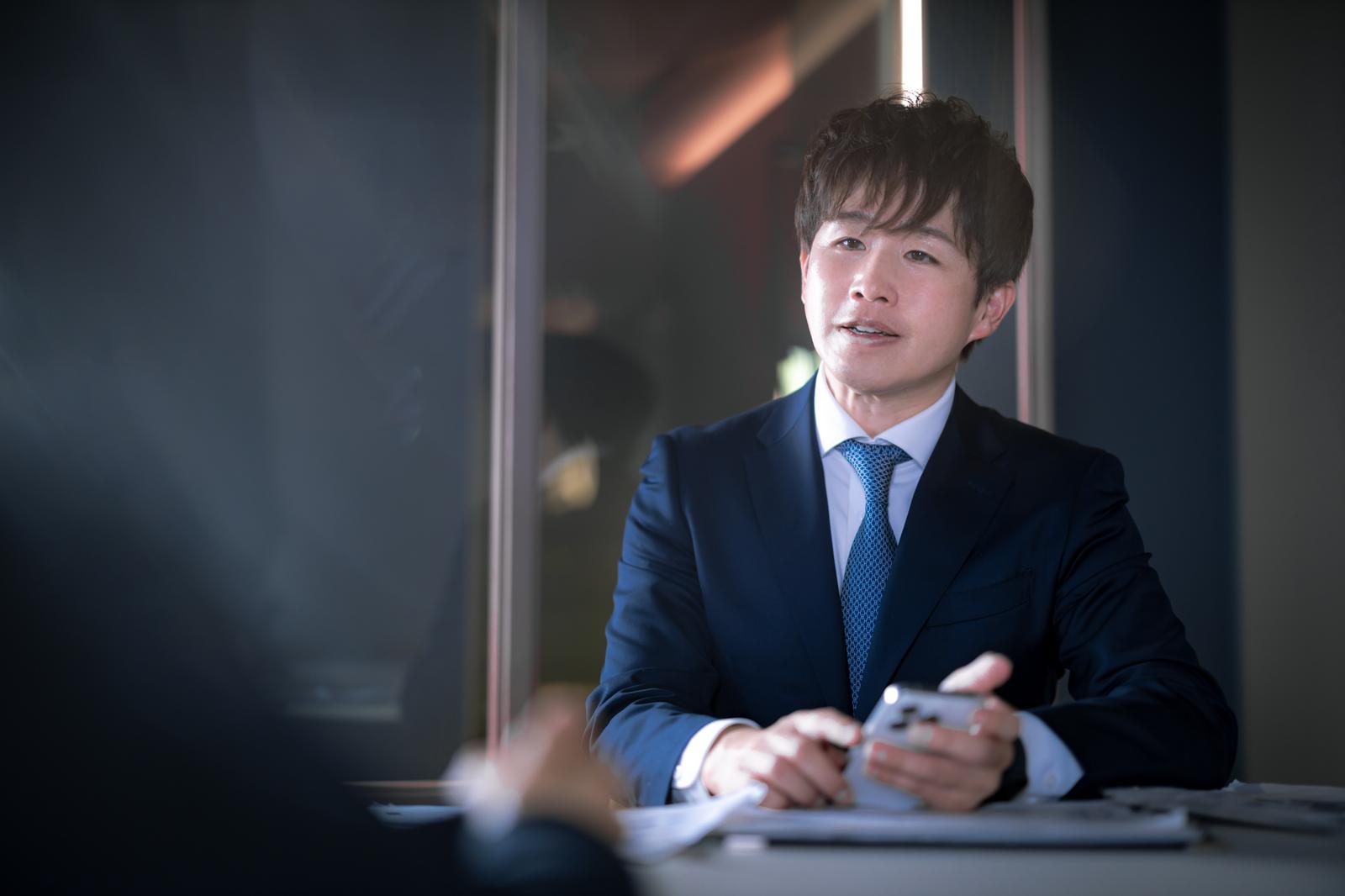 タブレットを持ってM&Aについて説明する紺色スーツの男性アドバイザー［モデル：野崎繁裕］