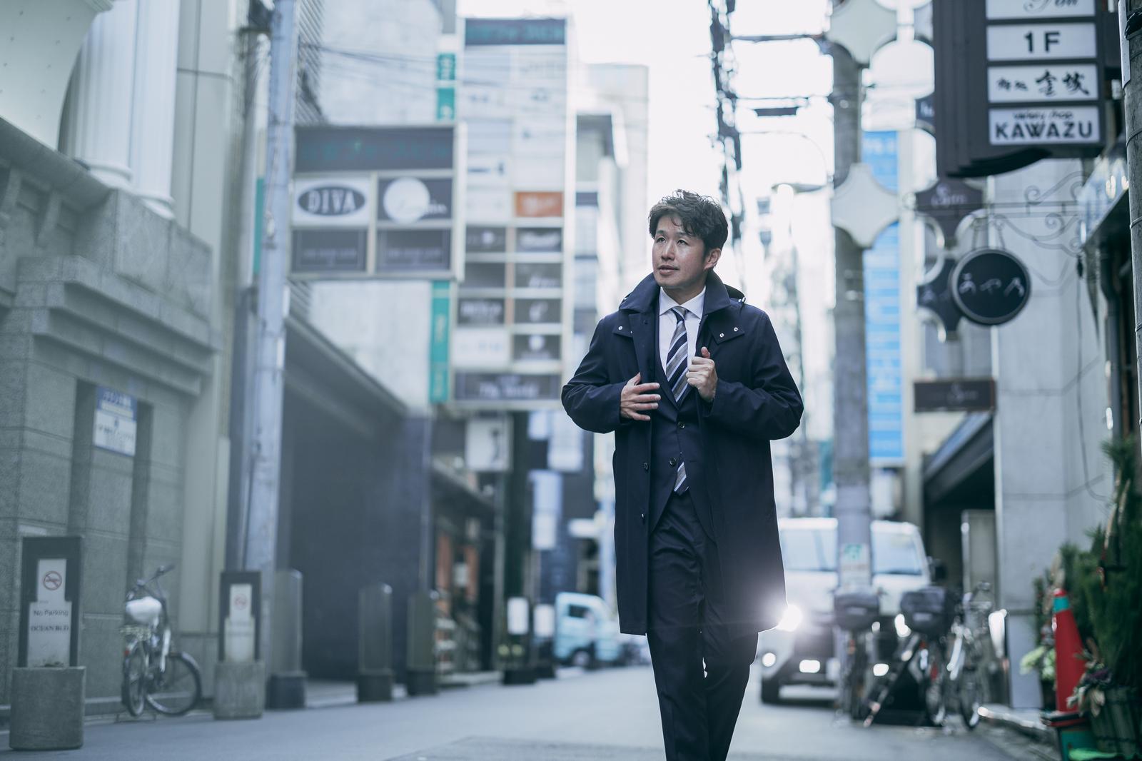 都市部の路地を歩くビジネススーツ姿の男性［モデル：野崎繁裕］
