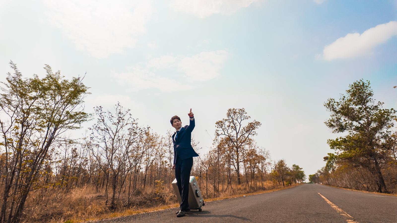 カンボジアの田舎道で親指を立ててヒッチハイクするスーツ姿の男性［モデル：野崎繁裕］