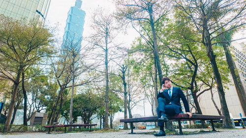 台北101と緑豊かな公園でベンチに座り電話する紺色スーツの男性