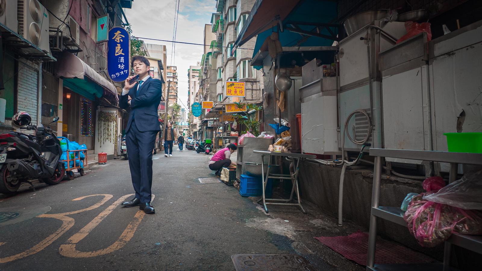 スーツケースを引くスーツ姿の男性が街路を歩く［モデル：野崎繁裕］