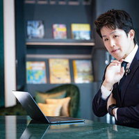 書棚のあるオフィスで頬杖をつき考え込むスーツ姿のビジネスマンの写真