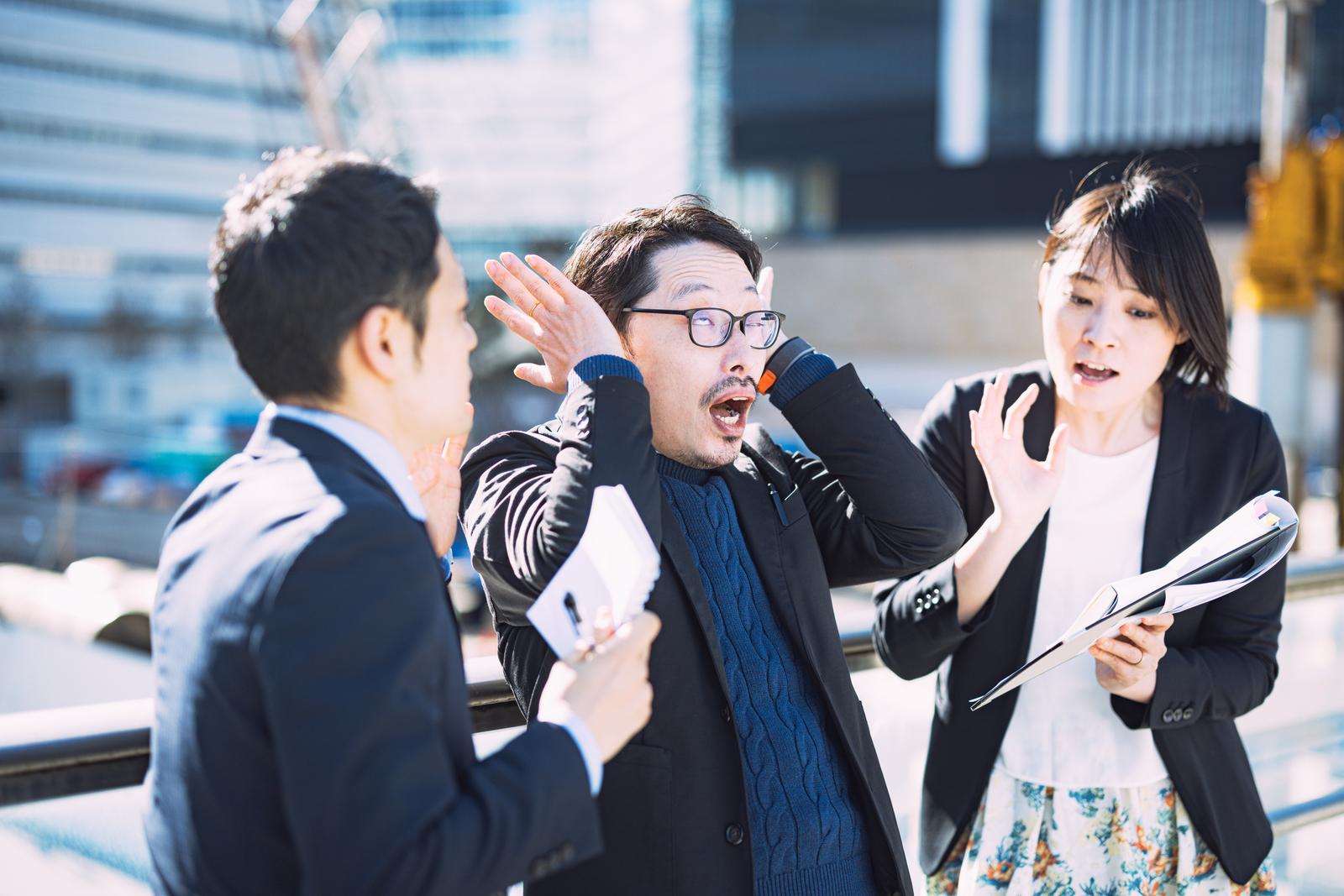 複数の質問に困惑して両手を広げる眼鏡をかけた男性上司と質問する男女社員［モデル：たけべともこ モデルリリース ゆうせい］
