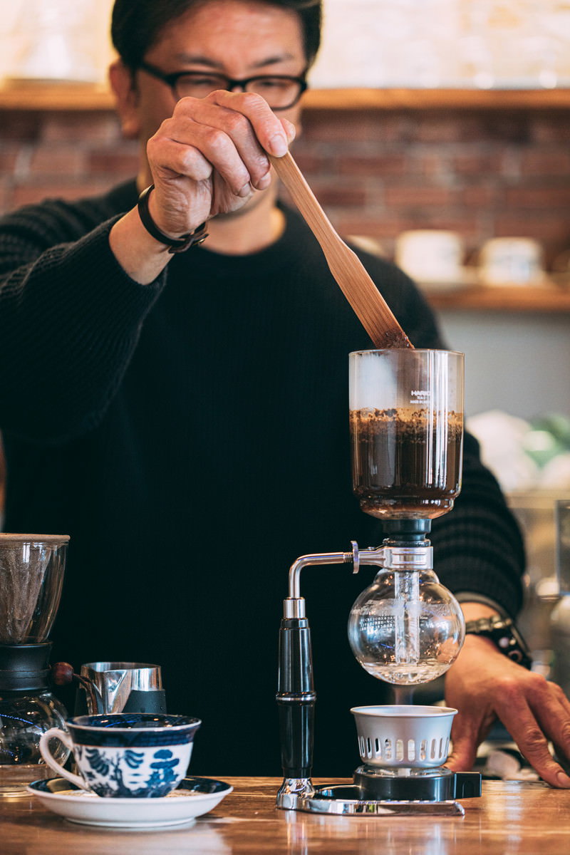 眼鏡をかけたカフェマスターがサイフォンを使ってコーヒーを抽出している様子