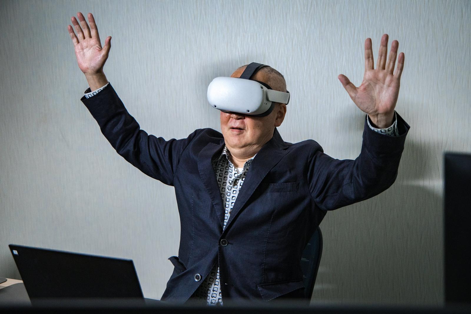 VRヘッドセットを装着して両手を上げているスーツ姿の男性［モデル：渡辺カンジ］