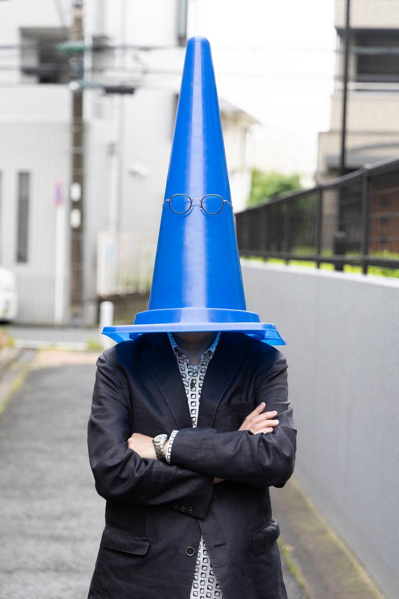 青い三角コーンを頭から被り腕組みをする黒ジャケットの男性［モデル：渡辺カンジ］