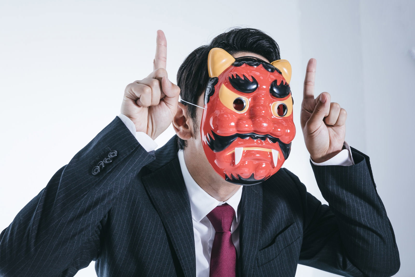 赤い鬼の面をつけて両手で指を立てて怒りを表現するスーツ姿の男性［モデル：大川竜弥］