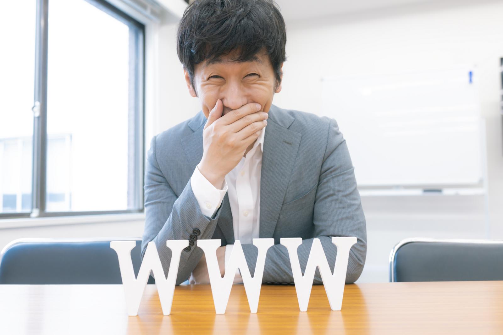 テーブルに「www」の文字を前に、口を手で押さえて笑う男性［モデル：大川竜弥］