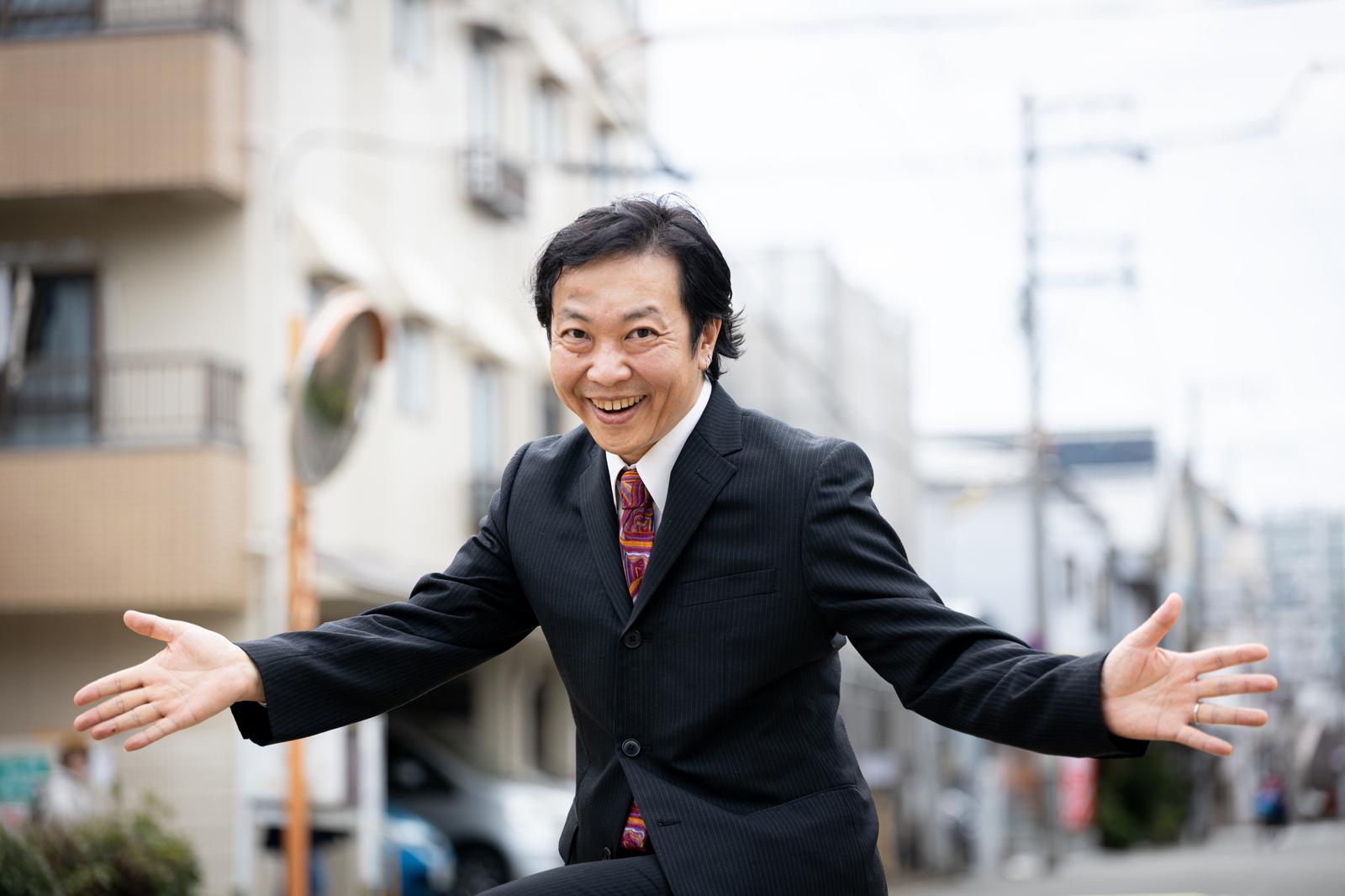 スーツ姿のおじさんが街中で両手を広げて笑顔を見せている［モデル：のせさん］