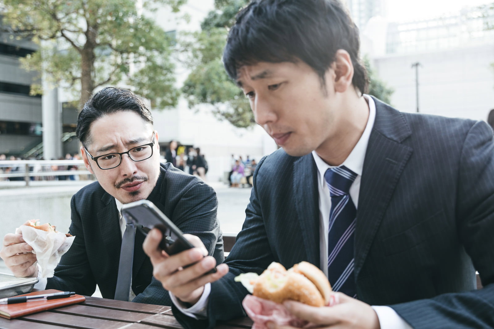 屋外でハンバーガーを食べながらスマホをのぞくスーツ男性2人［モデル：大川竜弥 ゆうせい］