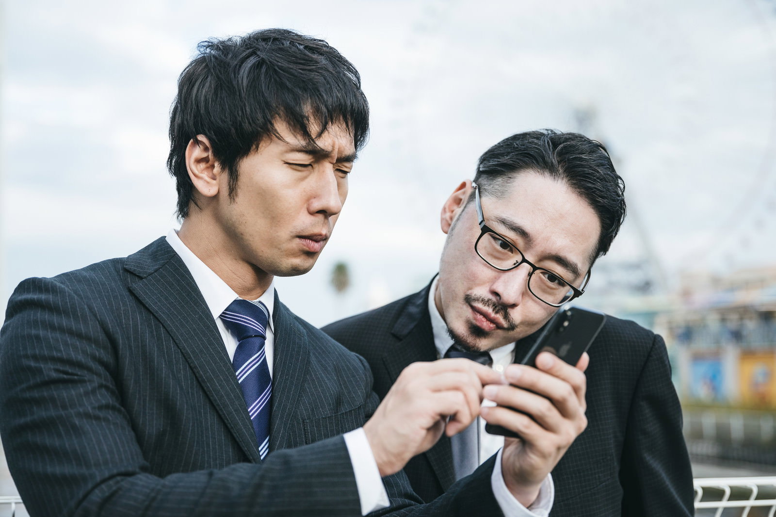 スーツを着た男性2人が屋外でスマートフォンを見ながらメモで相談している［モデル：大川竜弥 ゆうせい］