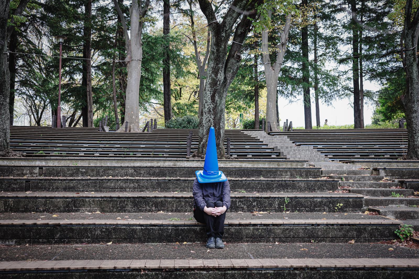 青いカラーコーンを頭にかぶって公園の階段にうつむいて座る人物