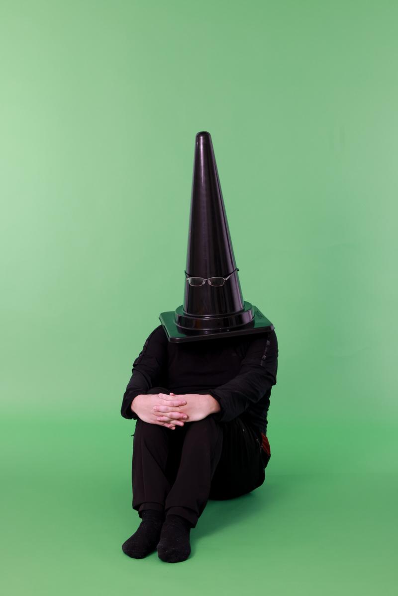 グリーンバックの前でカラーコーンを頭に被り体育座りをする人物