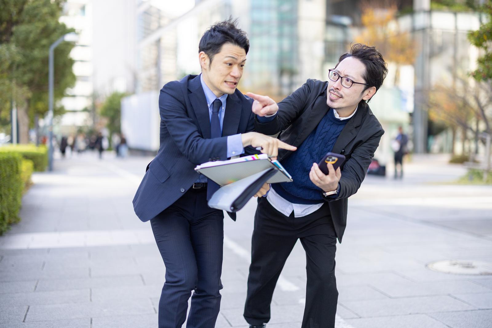 街中を移動しながら資料を確認する男性社員とスーツ姿の男性［モデル：ゆうせい モデルリリース］