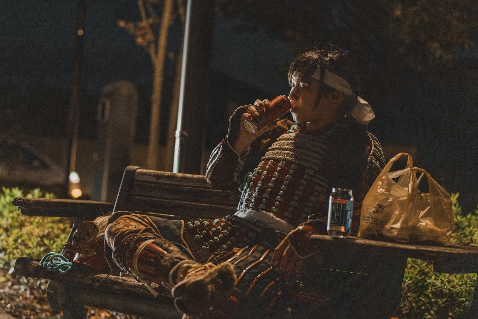 夜の公園のベンチで甲冑姿の男性がやけ酒をしている［モデル：まーこ］