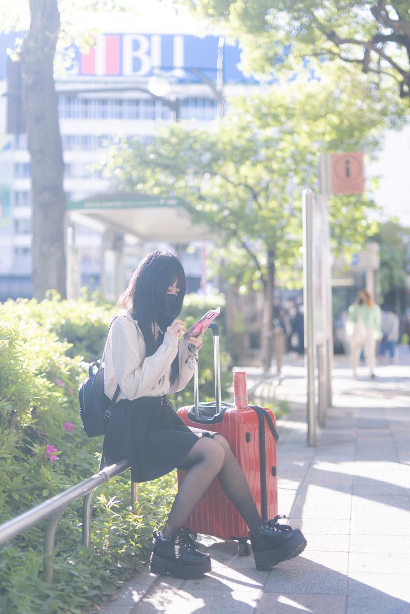 赤いキャリーケースとスマートフォンを持つマスク姿の女性が都会の駅前で待つ［モデル：緋真煉］